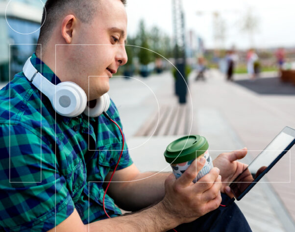 Man with headphones around his neck sits outside, holding a coffee cup and looking at a smartphone