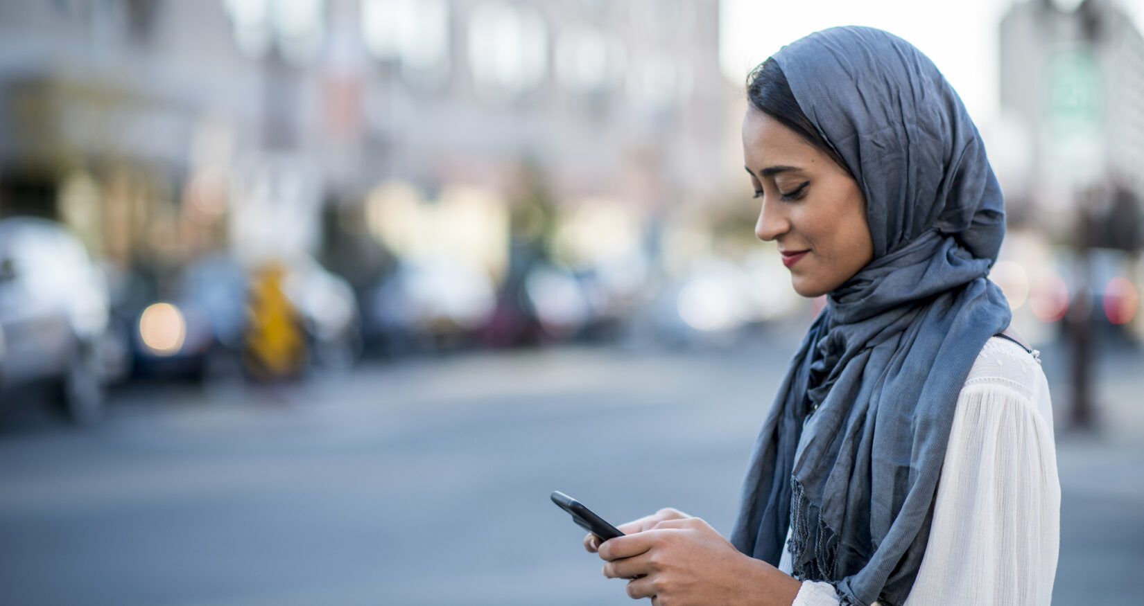 A Muslim woman is outdoors on a sunny day. She is wearing casual clothing and a head scarf. She is standing near a road and sending a message with her smartphone.